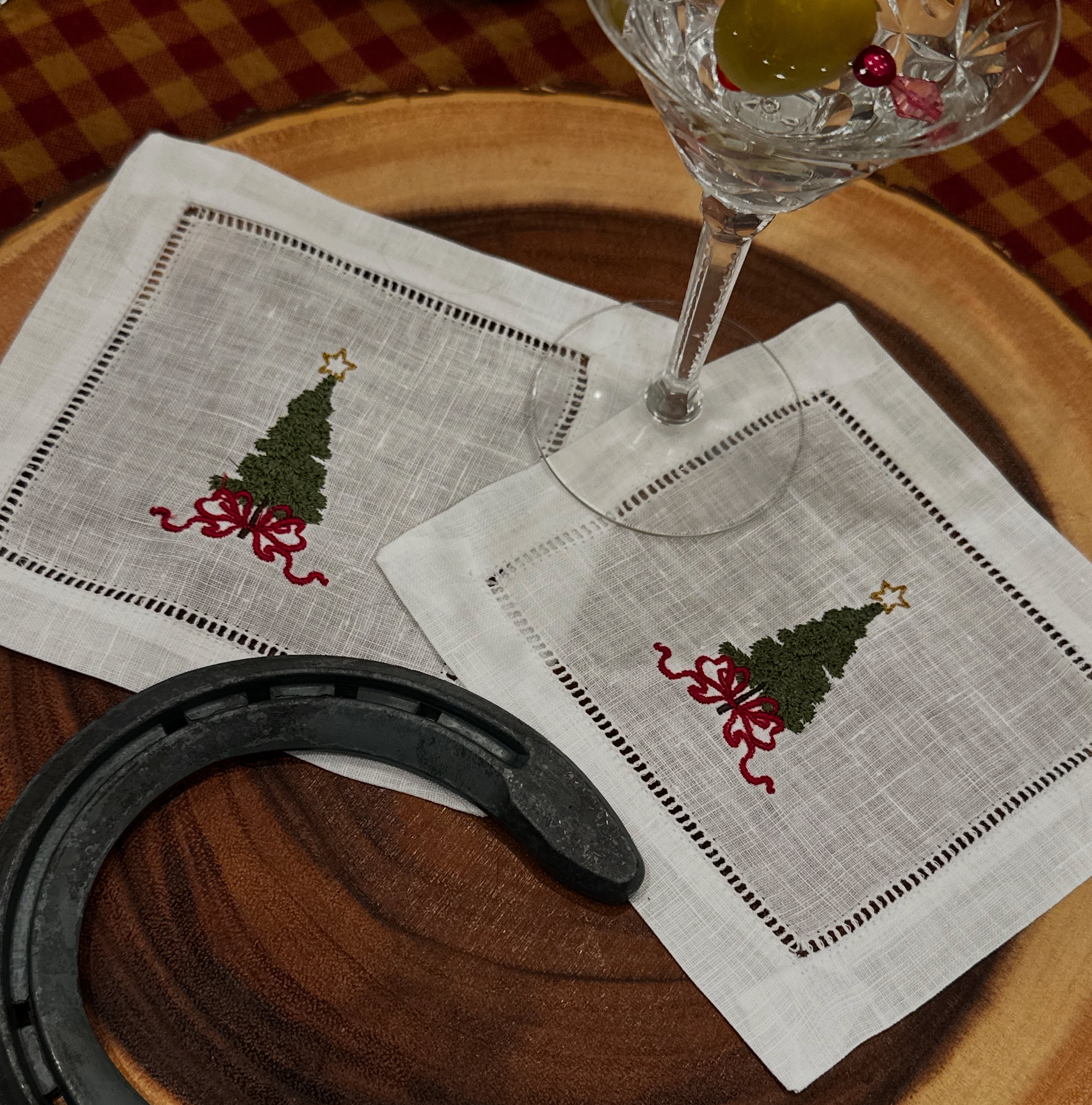 Two cocktail napkins with Christmas tree designs on a wooden table with a glass and horseshoe.