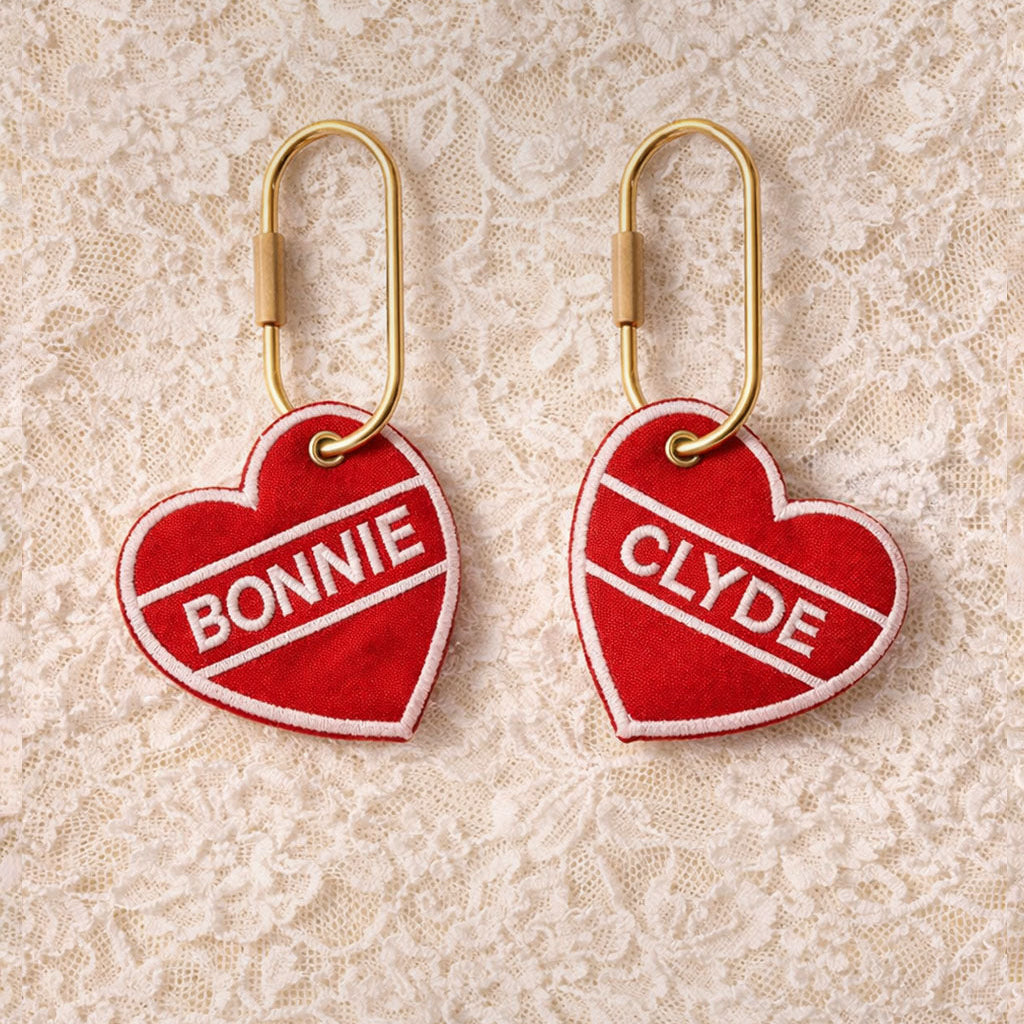 Two red heart-shaped keychains with 'Bonnie' and 'Clyde' text on a lace background