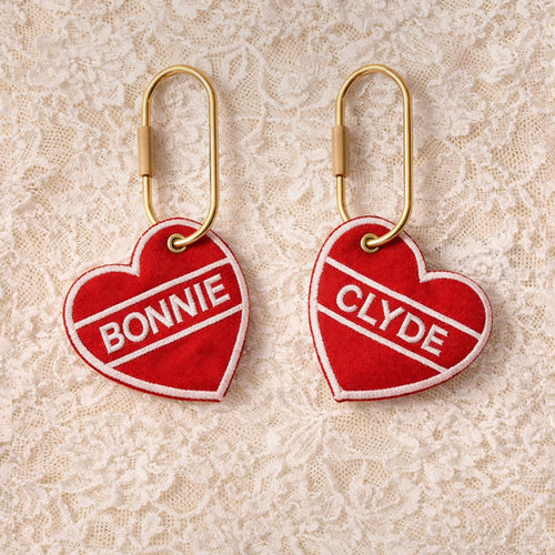 Two red heart-shaped keychains with 'Bonnie' and 'Clyde' text on a lace background